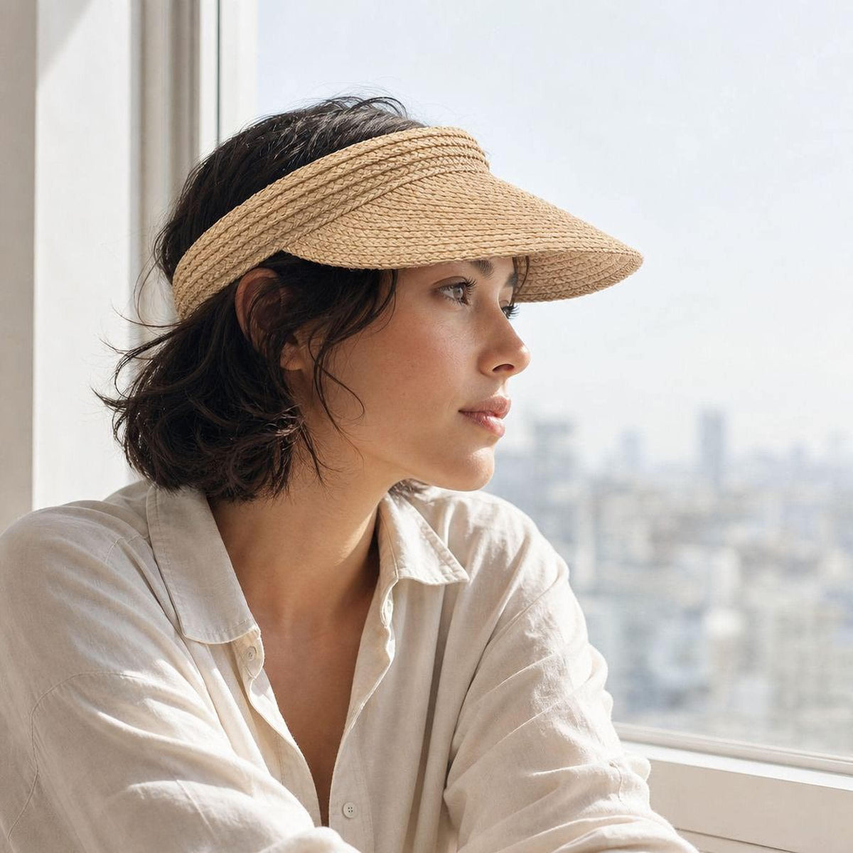 Woman wearing a handwoven raffia straw visor hat with open top, looking out a window over the city
