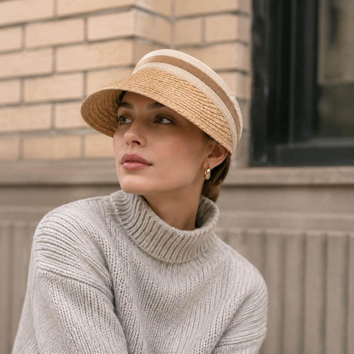 Handmade raffia straw sun visor with striped fabric headband, woman in beige sweater by a brick wall