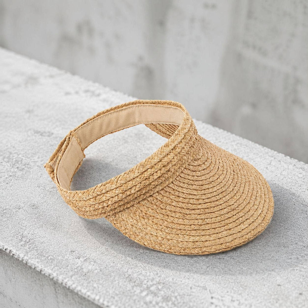 Packable handwoven raffia straw visor hat with adjustable closure on a concrete ledge